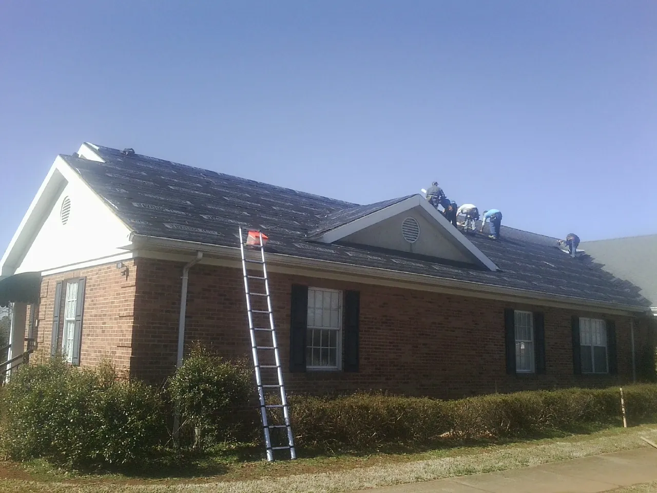 Crew tearing shingles off a brick ranch roof with a ladder set — Emergency Roof Repair work by our crew in Ortonville